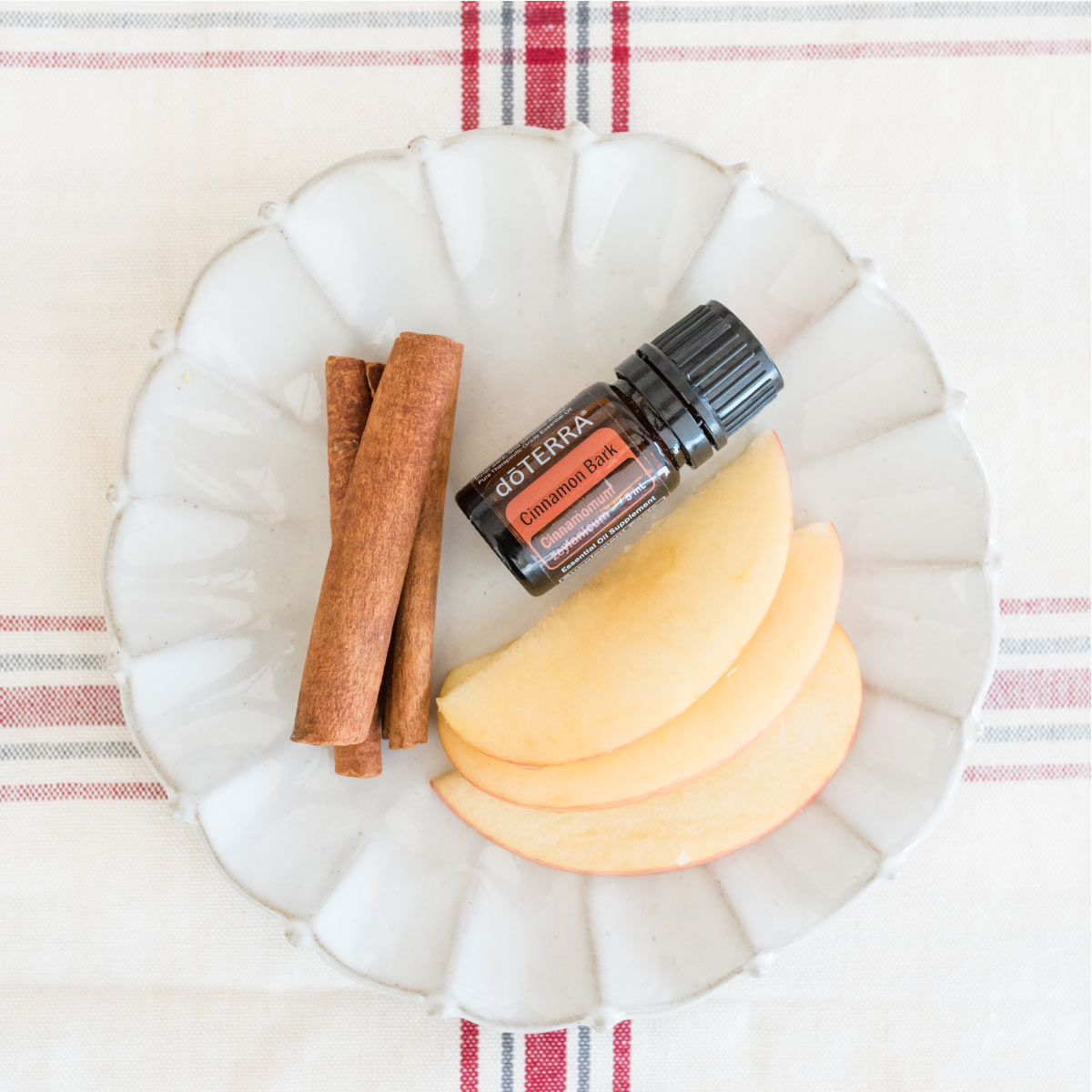 Bottle of Cinnamon essential oil on a plate with apple slices and fresh cinnamon spices. What are the common uses for Cinnamon Bark essential oil? doTERRA Cinnamon oil can be used to support a healthy immune system, for massage, for household cleaning, to add flavor to food, and more.