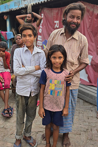 A man standing behind two children on a bustling street. His hand on the child’s shoulder and his other on the second child’s back.