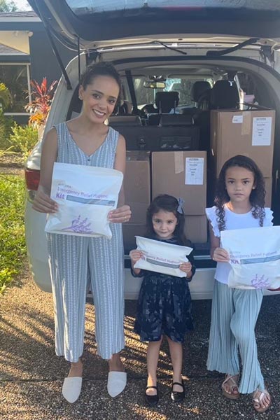 A woman and two young girls pose for a photo while holding doTERRA 72-Hour Emergency Relief Hygiene Kits. The trunk of an SUV is open and filled with brown boxes of hygiene kits.