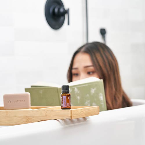  A woman sitting in a bathtub reading a book. doTERRA Serenity blend and Serenity Bath Bar sitting a wooden tray laying across the bathtub.