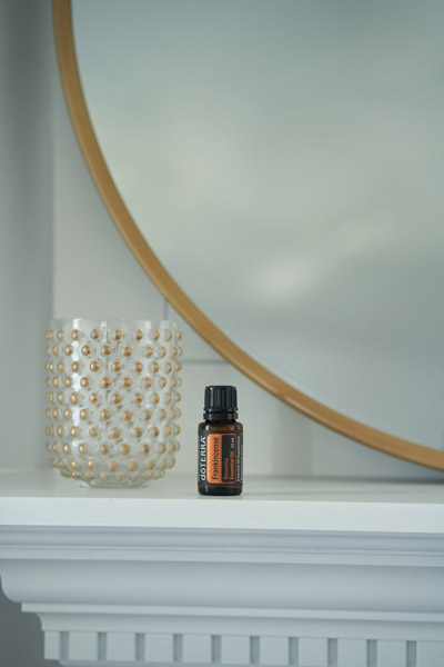 doTERRA Frankincense essential oil sitting on the edge of a white shelf. A round, gold mirror and a golden, dotted decoration sit behind the essential oil bottle.