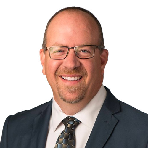 A professional headshot of Dr. Brannick Riggs, looking directly into the camera. Dr. Riggs is wearing a suit, tie, and white button-down shirt.