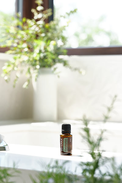 Bottle of doTERRA Eucalyptus essential oil sitting on the edge of a bathtub. Greenery in the front of the tub and a vase full of greenery sitting in the background.