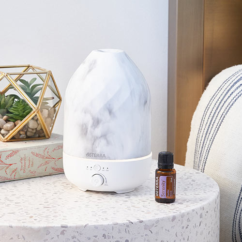 doTERRA Serenity blend sitting on a round nightstand table. The Volo Marble Diffuser sits beside it. A book and plant sit behind them both. 