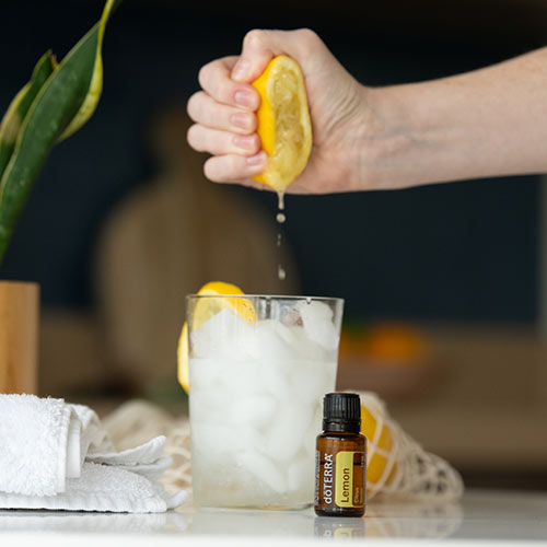 Someone squeezing a lemon into a glass of water and ice. doTERRA Lemon essential oil sits beside the glass.