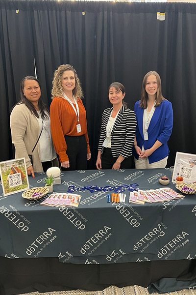 Four women stand behind a table with a tablecloth labeled ”doTERRA” in several places. Pens, pamphlets, a diffuser, and keychains lay across the table.