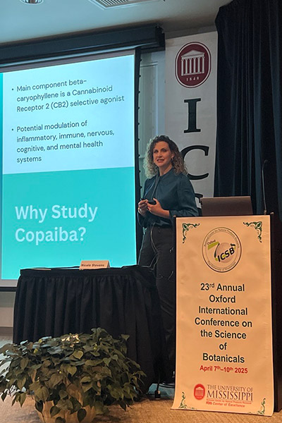Dr. Nicole Stevens stands on a stage presenting her studies on Copaiba. A screen showing her presentation is seen along with posters about ICSB. 