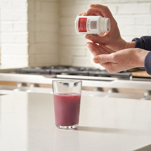 Person pouring doTERRA's GX Assist dietary supplement with glass of juice on a kitchen counter.
