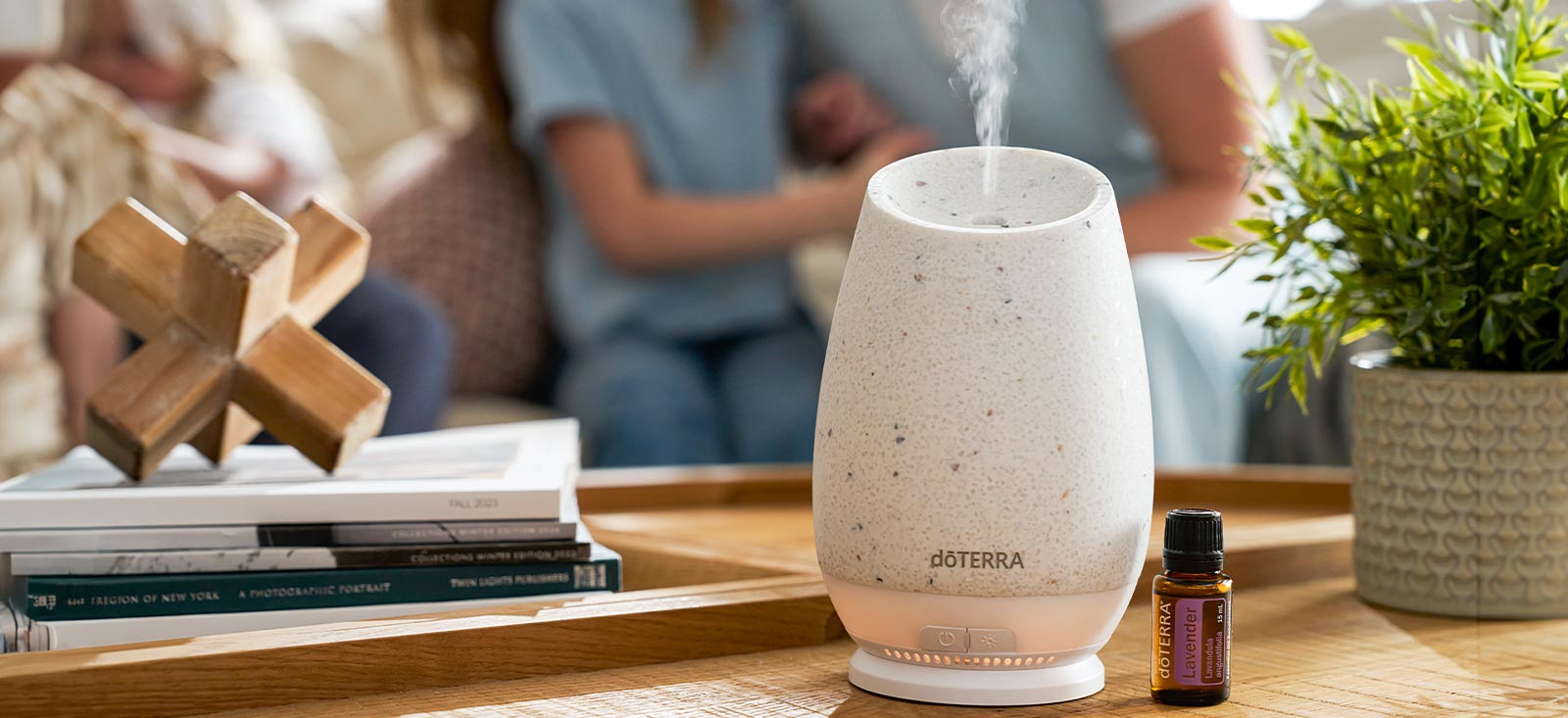 A close-up of a doTERRA Roam Diffuser and Lavender Essential Oil on a wooden table, and blurred people in the background.