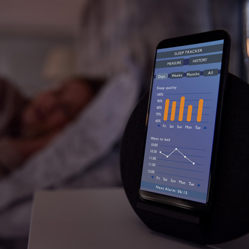 Smartphone displaying sleep tracker data on a nightstand with a person sleeping in the background.