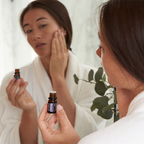 A woman in a white robe holding Tulsi Essential Oils and looking at the mirror.