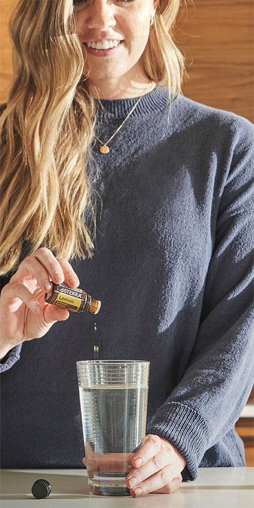 A woman with a bright smile pours doTERRA Lemon Essential Oil into a glass of water.
