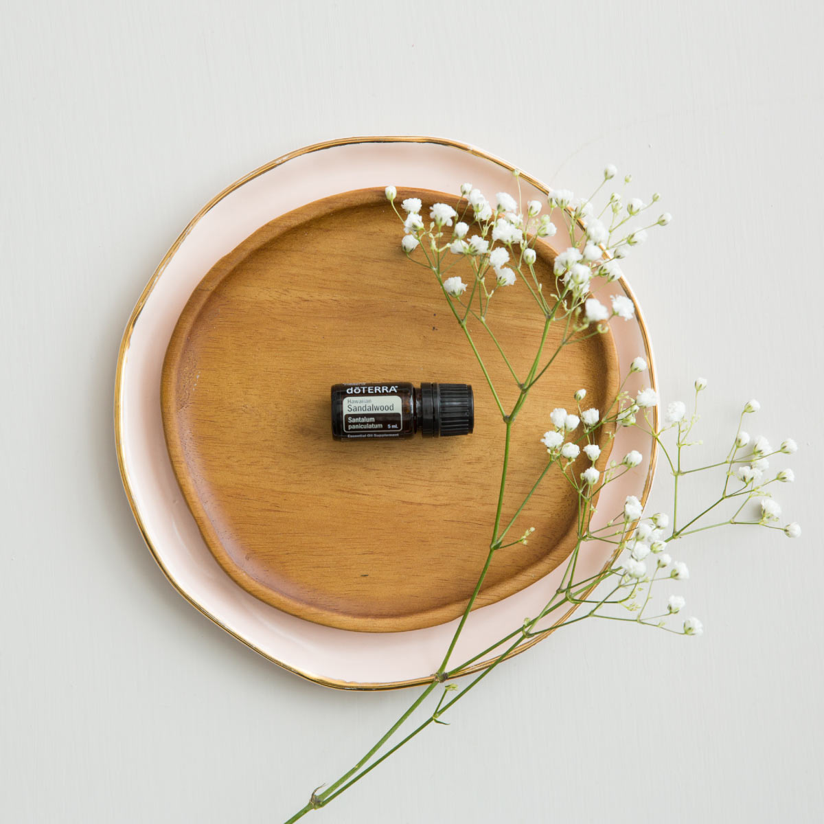 Bottle of doTERRA Hawaiian Sandalwood oil resting on a wooden dish next to small white flowers. There are dozens of benefits of Hawaiian Sandalwood oil, including benefits for skin, meditation, and promoting relaxation.